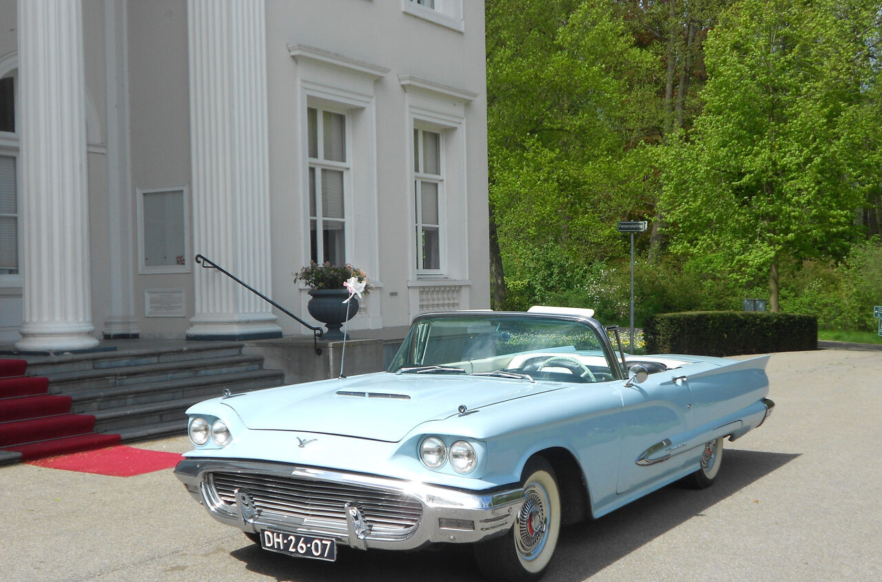 Poederblauwe Ford Thunderbird 1959 schuin van voren voor een wit landhuis met zuilen.
